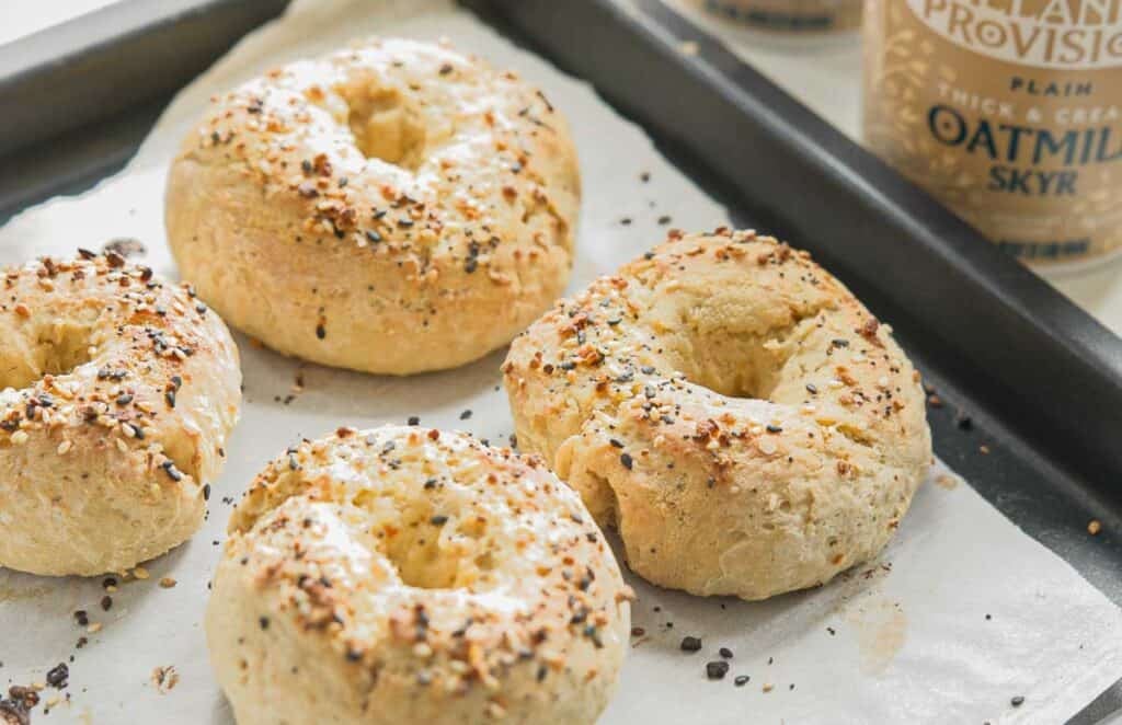 Four freshly baked bagels with seasoning on top are placed on a parchment-lined baking tray. A container of oatmilk skyr is visible in the background.